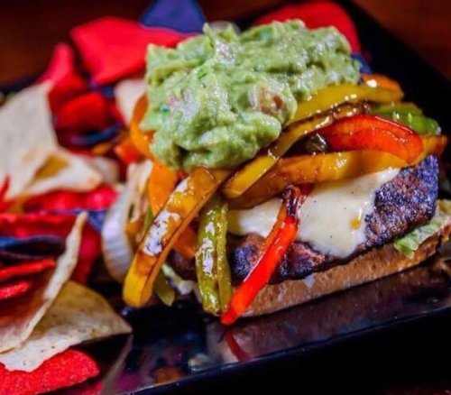 A delicious burger topped with guacamole and colorful sautéed peppers, served with tortilla chips on the side.