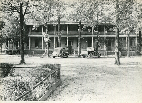 Black and white photo of a two-story building with cars parked in front, surrounded by trees and a grassy area.