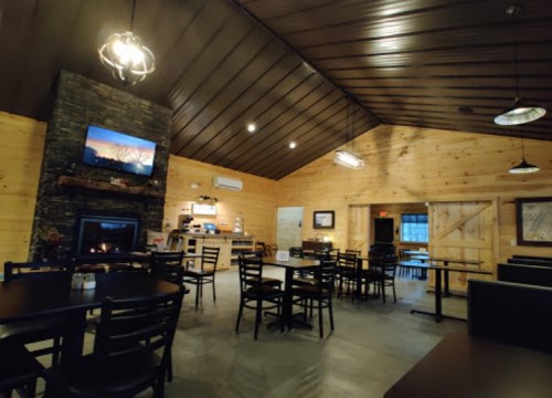 Cozy restaurant interior with wooden walls, a stone fireplace, and tables set for dining. Warm lighting creates a welcoming atmosphere.