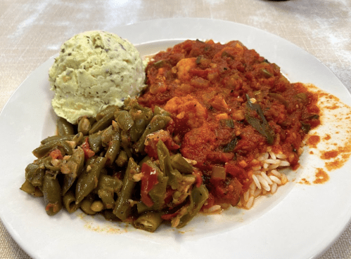 A plate of rice topped with shrimp in tomato sauce, served with green beans and a scoop of mashed potatoes.