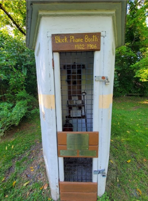 A vintage phone booth from 1902-1996, featuring a wooden sign and a metal grate, set in a green outdoor area.