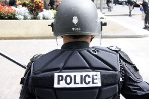 A police officer in tactical gear stands with their back to the camera, displaying a "POLICE" label on their uniform.
