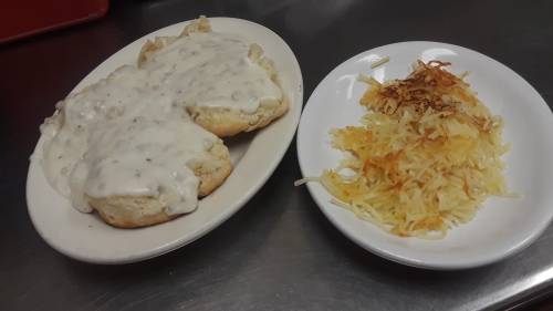 A plate with two biscuits smothered in creamy gravy, alongside a serving of crispy hash browns.