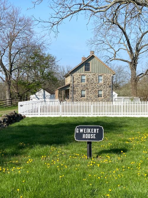 A stone house with a green roof, surrounded by a white picket fence and grassy yard, marked by a sign reading "G. Weikert House."