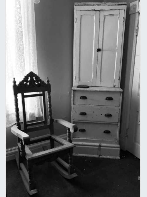 A vintage rocking chair next to a weathered cabinet with drawers in a softly lit room.