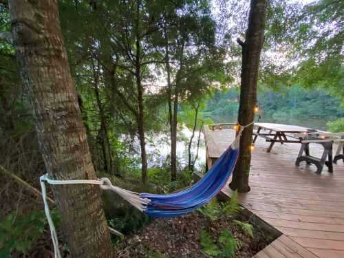 A colorful hammock hangs between two trees near a wooden deck overlooking a serene lake surrounded by greenery.