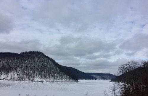 A snowy landscape with a frozen lake surrounded by hills under a cloudy sky.