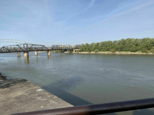 A calm river flows under a bridge, surrounded by green trees and a clear blue sky.