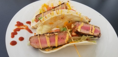 Three tacos filled with grilled steak, lettuce, tomatoes, and drizzled with sauce on a white plate.