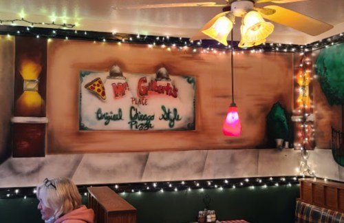 Cozy restaurant interior with a mural of "Mr. Gilbert's Place" and festive string lights. A woman sits at a table.