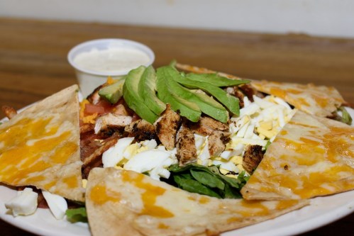 A plate of nachos topped with grilled chicken, avocado, diced tomatoes, cheese, and a side of dipping sauce.