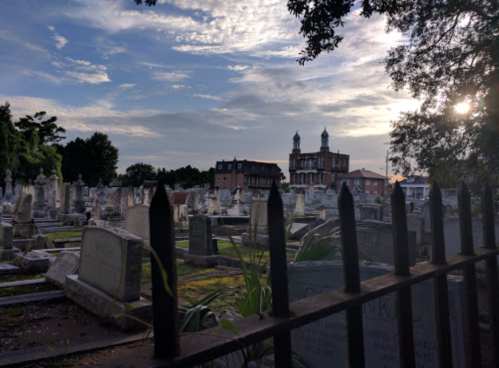 A cemetery with gravestones under a cloudy sky, featuring a historic building in the background and sunlight peeking through.