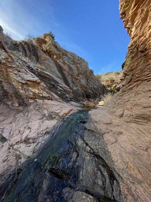 A narrow canyon with rocky walls and a small stream flowing through, under a clear blue sky.