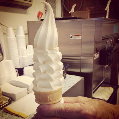 A hand holding a tall, swirled soft serve ice cream cone in a waffle cone, with a kitchen background.
