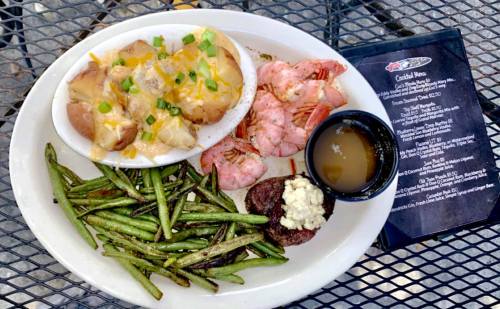A plate of shrimp, baked potatoes with cheese, green beans, and a small portion of steak, served with a menu on the side.