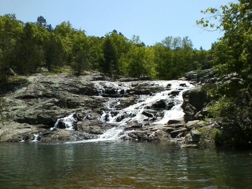 A serene waterfall cascades over rocky terrain, surrounded by lush green trees under a clear blue sky.