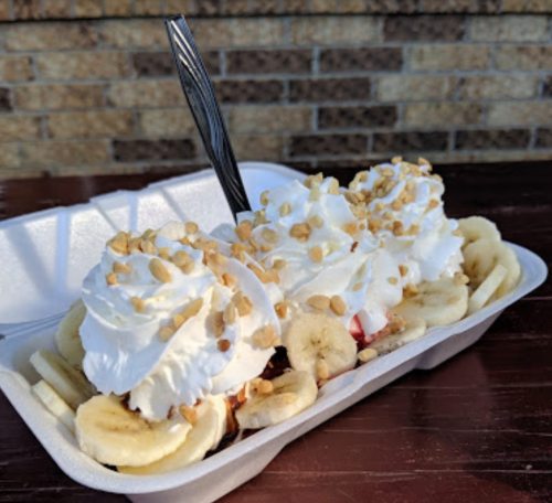 A dessert in a foam container featuring bananas, whipped cream, and chopped nuts.