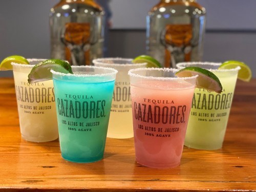 Five colorful cocktails in branded cups, garnished with lime, on a wooden table with tequila bottles in the background.