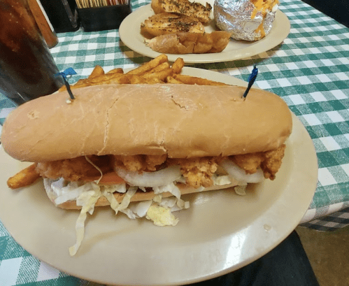 A large sandwich with fried chicken, lettuce, and onions, served with a side of fries on a checkered tablecloth.