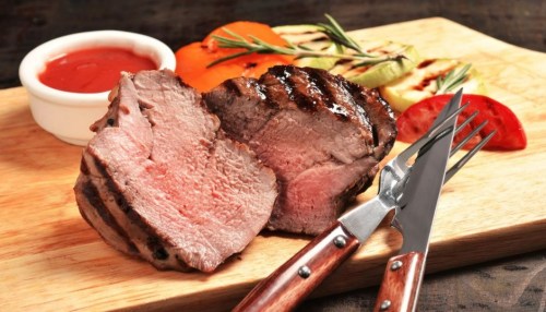 Grilled meat on a wooden board, accompanied by grilled vegetables and a small bowl of sauce. Fork and knife beside it.