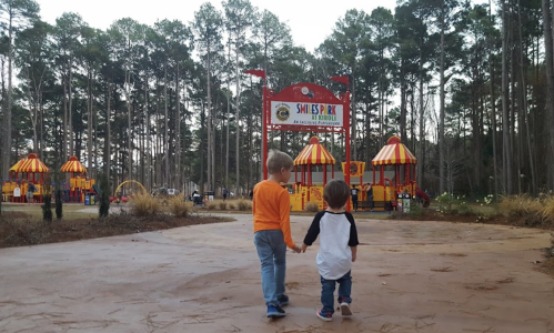 Two young children walk hand in hand towards a colorful playground surrounded by tall trees.