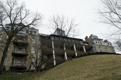 A large, historic building with multiple stories, set on a grassy hill, under a cloudy sky.