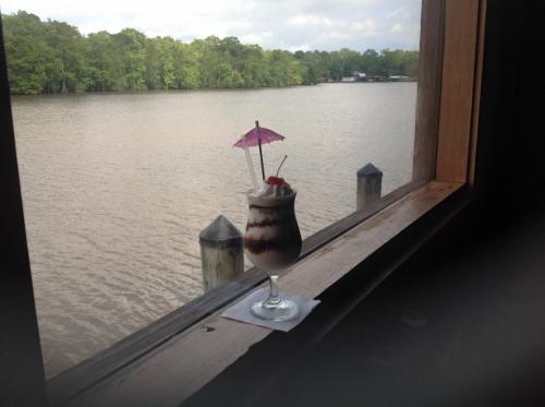 A tropical drink with an umbrella and cherry sits on a windowsill, overlooking a calm river and lush greenery.