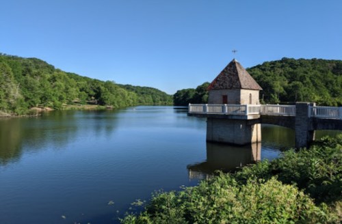 A serene lake scene featuring a small stone building on a pier, surrounded by lush green trees under a clear blue sky.