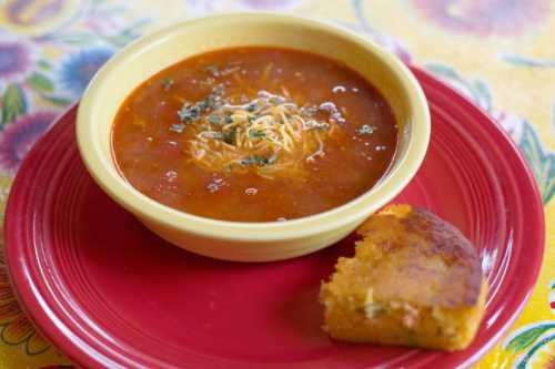 A bowl of soup with noodles and herbs, served on a red plate alongside a piece of cornbread.