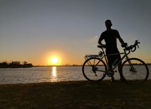Silhouette of a person with a bicycle at sunset by the water, with a warm orange glow in the sky.