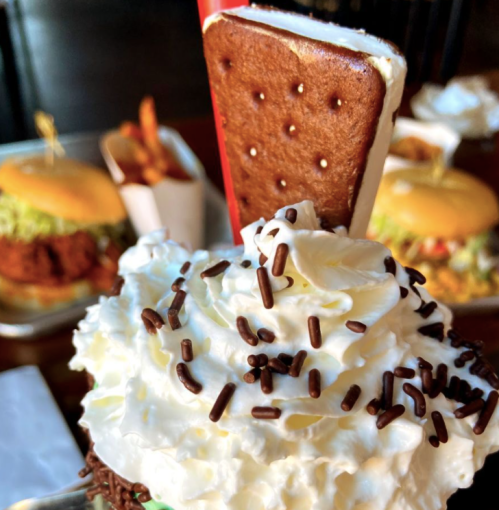 A close-up of a dessert topped with whipped cream, chocolate sprinkles, and a cookie, with burgers and fries in the background.