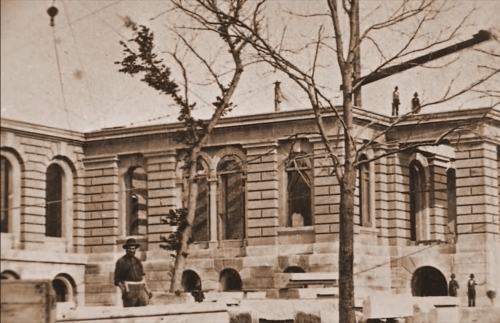 Historic building under construction, featuring stonework and large windows, with a tree and a worker in the foreground.