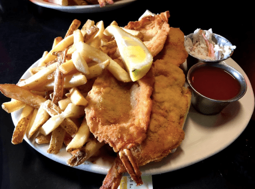 A plate of fried fish fillets with French fries, a lemon wedge, coleslaw, and dipping sauce.