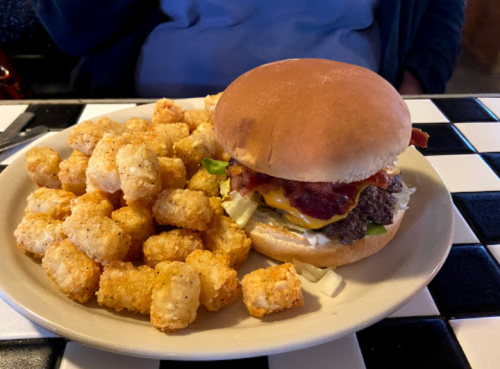 A plate with a cheeseburger topped with bacon and a side of crispy tater tots on a checkered table.