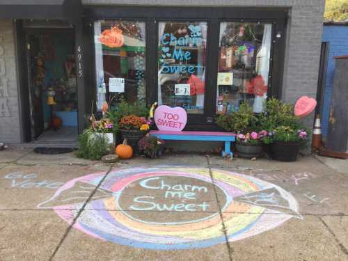 A colorful storefront with a chalk drawing on the sidewalk reading "Charm me Sweet" and vibrant flower pots.