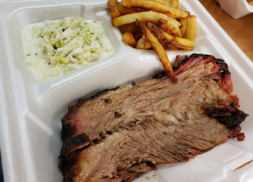 A serving of sliced brisket with coleslaw and French fries in a takeout container.