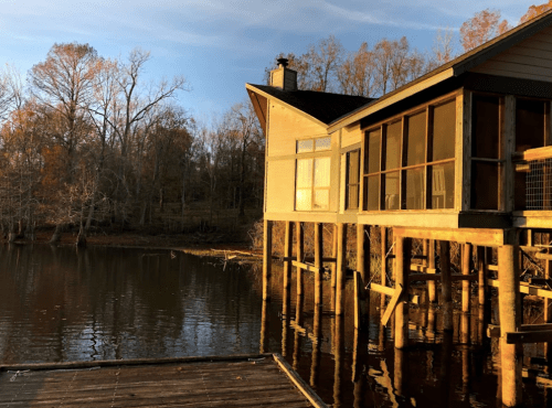 A modern house on stilts by a calm lake, surrounded by trees with autumn foliage under a clear sky.