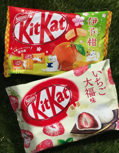 Two packages of Nestlé KitKat: one with orange flavor and the other with strawberry flavor, set on green grass.