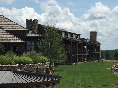 A rustic building with stone accents and balconies, surrounded by green grass and trees under a cloudy sky.