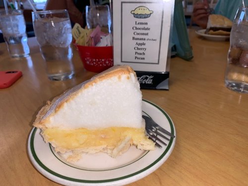 A slice of pie with a fluffy meringue topping on a plate, set on a wooden table with drinks in the background.