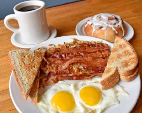 A plate with two fried eggs, crispy bacon, hash browns, and toast, alongside a cup of coffee and a cinnamon roll.