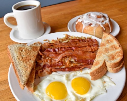 A plate with two fried eggs, crispy bacon, hash browns, and toast, alongside a cup of coffee and a cinnamon roll.