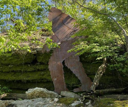 A large metal silhouette of a Bigfoot figure stands among trees and rocks in a natural setting.