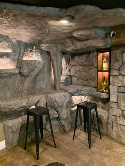 A rustic bar area with rock-like walls, featuring two black stools and shelves displaying bottles.