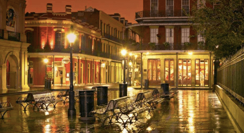 A quiet, illuminated street in a historic district, with benches and wet pavement reflecting warm lights at dusk.