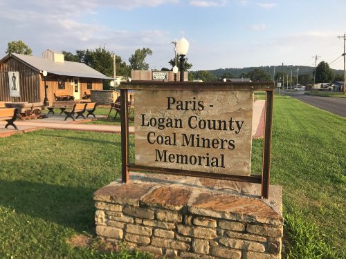 Sign for the Paris-Logan County Coal Miners Memorial, with a grassy area and buildings in the background.