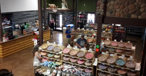A spacious store interior with wooden displays filled with colorful candies and snacks, and customers browsing around.