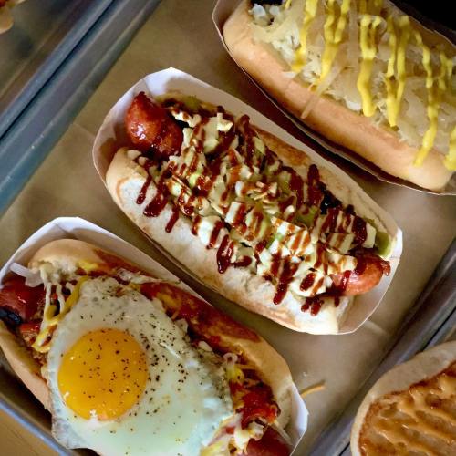 A variety of gourmet hot dogs, including one topped with an egg, drizzled sauces, and fresh toppings in paper trays.