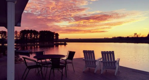 A serene lakeside view at sunset, featuring a table and chairs on a deck with colorful clouds reflecting on the water.