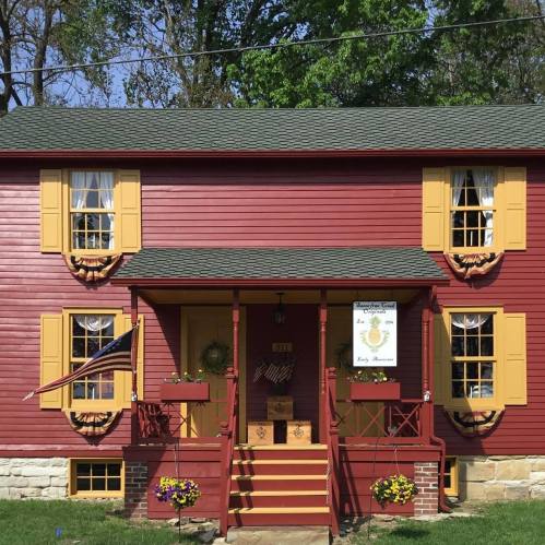 A charming red house with yellow shutters, a welcoming front porch, and flower beds, surrounded by greenery.